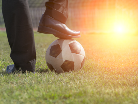 close-up of a person’s foot resting on a soccer ball on a grassy field, with sunlight in the background.
