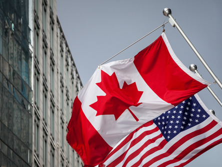 The Canadian and American flags are flying side by side on flagpoles outside a modern building.