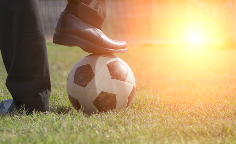 close-up of a person’s foot resting on a soccer ball on a grassy field, with sunlight in the background.