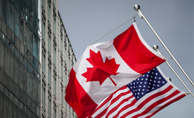 The Canadian and American flags are flying side by side on flagpoles outside a modern building.