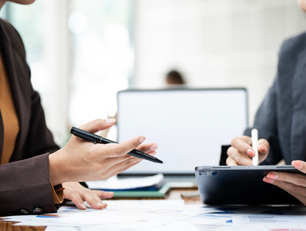 Two people in business attire discussing documents; one holds a pen and paper, the other uses a stylus on a tablet. A laptop is open on the desk in the background.