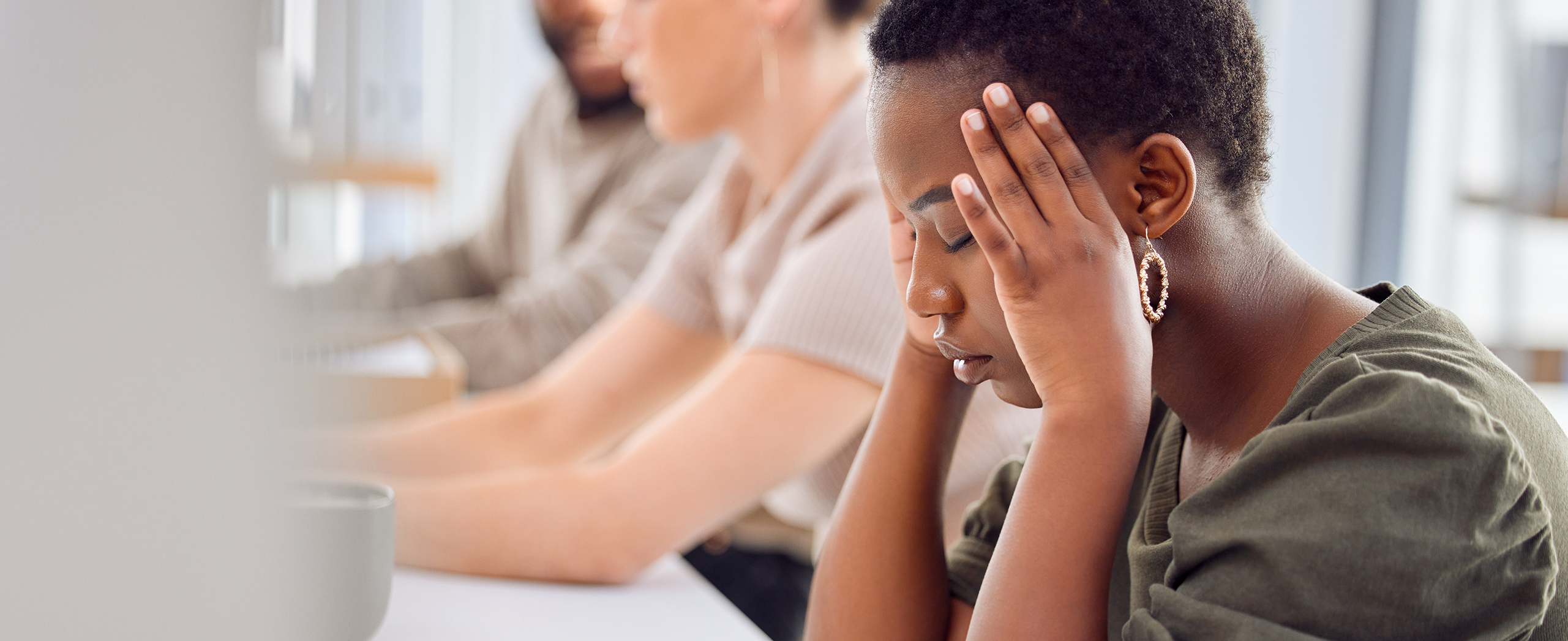 A Black woman sits at a desk holding her head in their hands, appearing stressed, with two colleagues in the background.