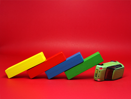 A toy bus is tipped over with four colored blocks—yellow, red, blue, and green—stacked and falling like dominoes against it, set on a red background.