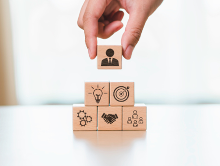 A hand places a wooden block with a person icon on top of a pyramid of blocks depicting business and teamwork symbols.