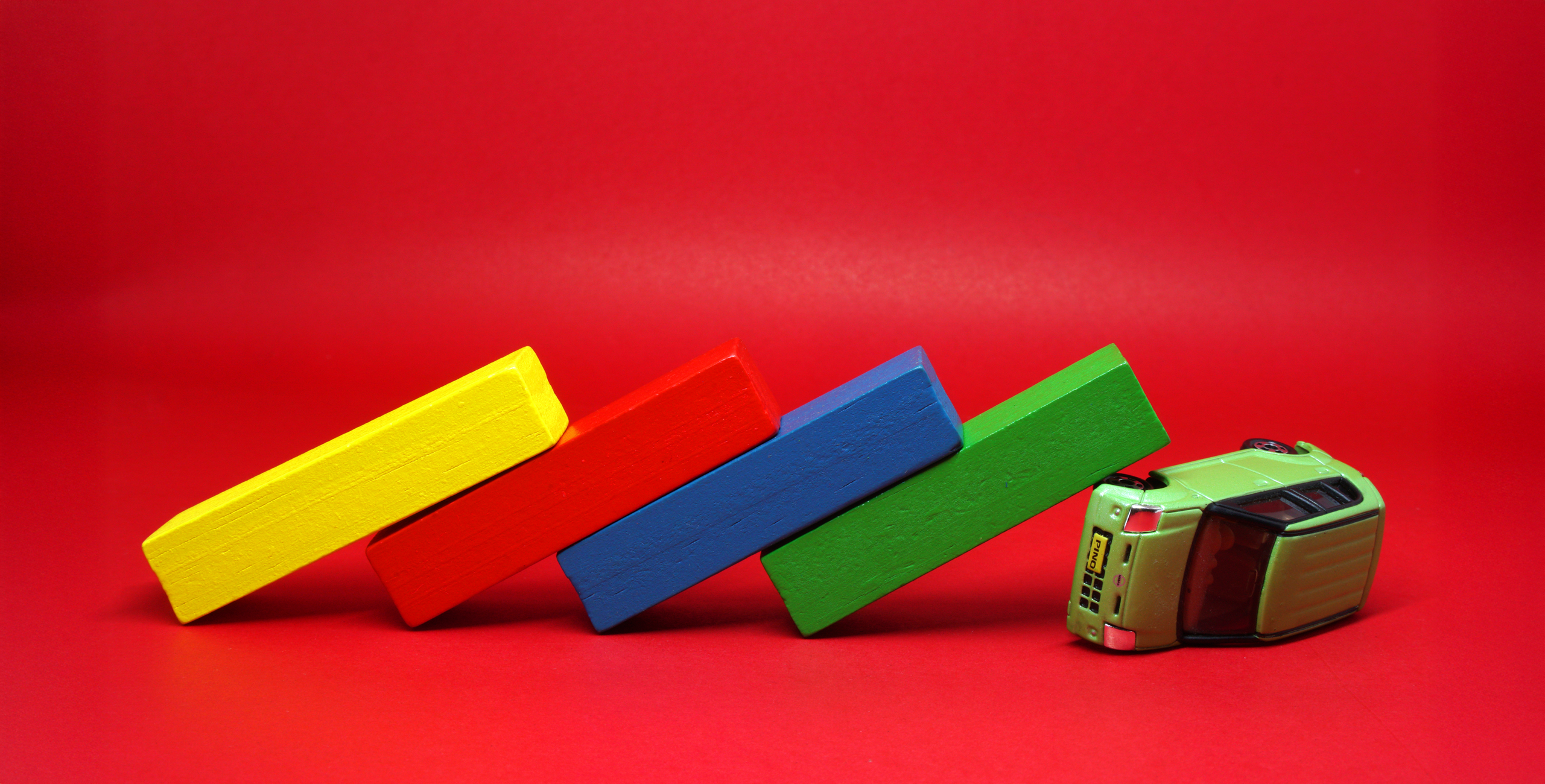 A toy bus is tipped over with four colored blocks—yellow, red, blue, and green—stacked and falling like dominoes against it, set on a red background.