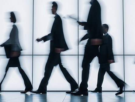 Blurred silhouettes of business professionals walking past a glass wall, suggesting movement and anonymity