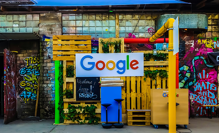 A decorated area with a large Google sign, wooden pallets, green plants, and graffiti-covered walls in the background.