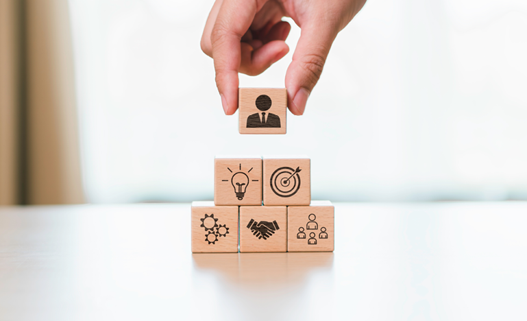 A hand places a wooden block with a person icon on top of a pyramid of blocks depicting business and teamwork symbols.