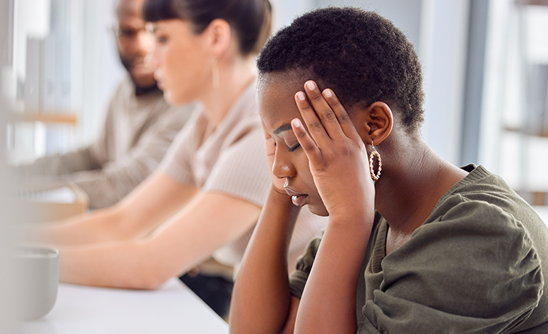 A Black woman sits at a desk holding her head in their hands, appearing stressed, with two colleagues in the background.