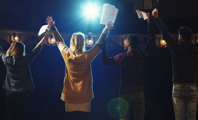 A group of people standing outdoors at night with their arms raised in celebration, illuminated by bright stage lights.