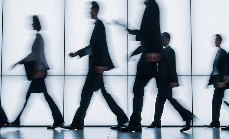 Blurred silhouettes of business professionals walking past a glass wall, suggesting movement and anonymity