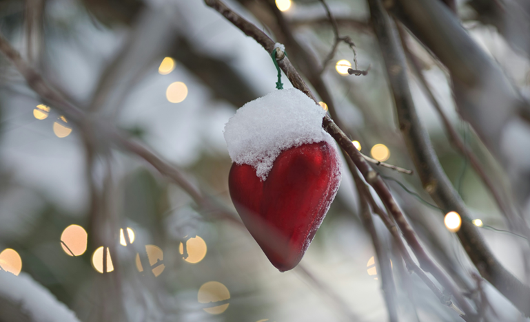 A red heart-shaped ornament dusted with snow hangs from a tree branch, surrounded by blurred string lights in the background.